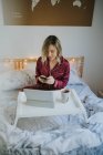Joven mujer bonita en pijama usando teléfono inteligente mientras está sentado en la cama con café y tableta en la bandeja de la mesa - foto de stock