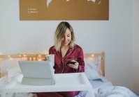 Молода красива жінка в піжамі використовує смартфон, сидячи в ліжку з кавою і планшетом на столі — стокове фото