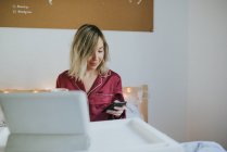 Mujer bonita joven en pijama usando teléfono inteligente mientras está sentada en la cama con bandeja de café y mesa - foto de stock