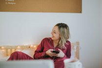 Joven mujer riendo en pijama sosteniendo un teléfono inteligente mientras está sentada en la cama - foto de stock