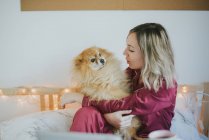 Junge glücklich lächelnde attraktive Frau im Pyjama sitzt im Bett mit kleinem flauschigen Hund — Stockfoto