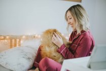 Young happy smiling attractive woman in pajamas sitting in bed with little fluffy dog — Stock Photo