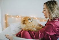 Jeune femme heureuse et souriante en pyjama assise au lit avec un petit chien moelleux — Photo de stock
