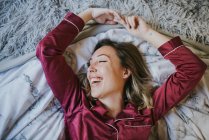 Cheerful young pretty woman in pajamas smiling with closed eyes on bed in bedroom — Stock Photo