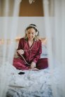 Молода щаслива усміхнена жінка в піжамі з навушниками та смартфонами сидить на ліжку вранці — стокове фото
