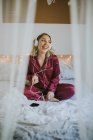 Молода щаслива усміхнена жінка в піжамі з навушниками та смартфонами сидить на ліжку вранці — стокове фото