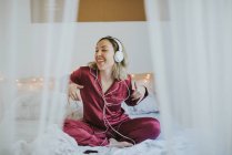 Young happy smiling woman in pajamas with headphones and smartphone listening to music in bed in morning — Stock Photo