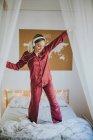 Молодая счастливая улыбающаяся женщина в пижаме с наушниками, танцующими на кровати утром — стоковое фото