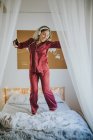 Молода щаслива усміхнена жінка в піжамі з навушниками танцює на ліжку вранці — стокове фото