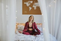 Молода жінка в піжамах медитує, сидячи в позі лотоса на зручному ліжку вранці — стокове фото