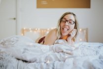 Jovem mulher alegre em óculos e roupa interior deitada na cama de manhã — Fotografia de Stock
