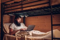 Концентрований бородатий хіпстер працює на ноутбуці, лежачи в простому двоярусному ліжку заміського дерев'яного будинку — стокове фото