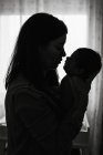 Silhueta de jovem mãe segurando em mãos bebê recém-nascido enquanto em pé em casa — Fotografia de Stock