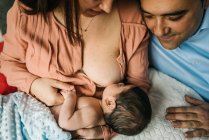 Зверху безликі мати і тато тримаються за руки і годують новонароджену дитину, загорнуту в ковдру на ліжку вдома — стокове фото