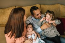 Felice giovane madre che tiene il neonato avvolto in una coperta e padre che tiene la figlia seduta sul divano a casa — Foto stock