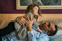 Pai se divertindo com a filha feliz segurando as mãos enquanto deitado na cama — Fotografia de Stock