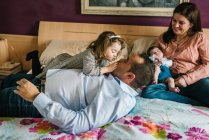 Père souriant tenant la petite fille riante couchée sur le lit avec la mère tenant le nouveau-né en arrière-plan dans la chambre — Photo de stock