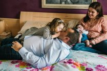 Улыбающийся отец, держащий смеющуюся маленькую дочь, лежащую на кровати с матерью, держащей новорожденного ребенка на заднем плане в спальне — стоковое фото