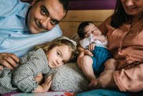 Padre sorridente che sorride piccola figlia sdraiata sul letto con madre che tiene il neonato sullo sfondo in camera da letto — Foto stock