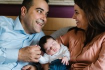 Sorrindo pai segurando rindo pequena filha deitada na cama com a mãe segurando bebê recém-nascido — Fotografia de Stock