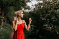 Боковой вид привлекательной блондинки, одетой в красную чувственную позу и трогающей шею с закрытыми глазами среди зеленых цветущих деревьев — стоковое фото