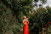 Vista laterale di attraente bionda vestita di rosso sensualmente in posa con gli occhi chiusi tra alberi verdi in fiore — Foto stock