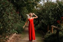 Vista lateral de la atractiva rubia vestida de rojo posando sensualmente con los ojos cerrados entre los árboles florecientes verdes - foto de stock