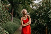 Vue latérale d'une jolie blonde vêtue de rouge posant sensuellement les yeux fermés parmi les arbres à fleurs vertes — Photo de stock
