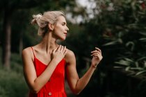 Side view of attractive blonde dressed in red sensually posing and touching her neck with closed eyes among green blooming trees — Stock Photo