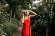 Vista lateral de loira atraente vestida de vermelho sensualmente posando com olhos fechados entre árvores verdes florescendo — Fotografia de Stock