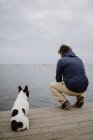 Back view of adult male and spotted French Bulldog sitting on timber pier and looking at calm sea on gray day — Stock Photo