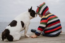 Visão traseira do garoto irreconhecível em jaqueta listrada batendo manchado Bulldog francês enquanto sentado no cais de madeira perto do mar — Fotografia de Stock