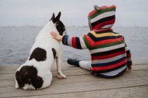 Vista trasera de niño irreconocible en chaqueta a rayas palmadas manchado Bulldog francés mientras está sentado en el muelle de madera cerca del mar - foto de stock