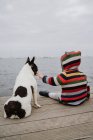 Vista trasera de niño irreconocible en chaqueta a rayas palmadas manchado Bulldog francés mientras está sentado en el muelle de madera cerca del mar - foto de stock