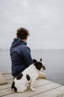 Hombre adulto anónimo con chaqueta cálida abrazando a Bulldog francés manchado mientras está sentado en el muelle de madera y admirando la vista del mar ondulante en un día aburrido - foto de stock