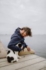 Adulto macho em casaco quente abraçando manchado Bulldog francês enquanto sentado no cais de madeira e admirando vista do mar ondulante no dia maçante — Fotografia de Stock