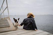 Vista posterior de la niña con Bulldog francés sentado en el muelle cerca del mar en el día nublado aburrido - foto de stock