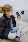Bambina con Bulldog francese seduta sul molo vicino al mare in una giornata opaca e nuvolosa — Foto stock