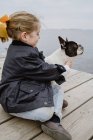 Bambina con Bulldog francese seduta sul molo vicino al mare in una giornata opaca e nuvolosa — Foto stock