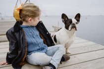 Petite fille avec Bulldog français assis sur la jetée près de la mer par une journée nuageuse terne — Photo de stock