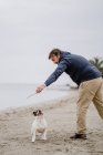 Vista lateral del hombre adulto con palo jugando con Bulldog francés obediente mientras pasa tiempo en la orilla arenosa cerca del mar - foto de stock