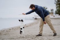 Vue latérale de l'homme adulte avec bâton jouant avec bouledogue français obéissant tout en passant du temps sur le rivage sablonneux près de la mer — Photo de stock