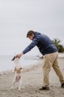 Side view of adult man with stick playing with obedient French Bulldog while spending time on sandy shore near sea — Stock Photo