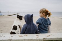 Задний вид на двух детей, обнимающих французский бульдог, сидящих на пляже рядом с морем вместе — стоковое фото
