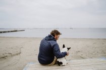 Hombre adulto con chaqueta abrigada abrazando a Bulldog francés manchado mientras está sentado en el muelle de madera y admirando la vista del mar ondulante en un día aburrido - foto de stock