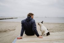 Erwachsene Männchen in warmer Jacke umarmen gefleckte Französische Bulldogge, während sie auf einem hölzernen Steg sitzen und den Blick auf das plätschernde Meer an einem trüben Tag bewundern — Stockfoto