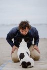 Uomo adulto in giacca calda abbracciando macchiato Bulldog francese mentre seduto sul molo di legno e ammirando vista del mare increspato il giorno noioso — Foto stock