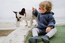 Entzückendes Kind lächelt und spielt mit Sonnenbrille, während es am Strand in der Nähe der französischen Bulldogge sitzt — Stockfoto
