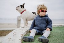 Приваблива дитина посміхається і бавиться з сонцезахисними окулярами, сидячи на пляжі біля пляжу французького бульдога — стокове фото
