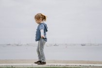 Вид збоку маленької дівчинки з простягнутими руками, що йдуть на кордоні з спокійним морем і сірим небом — стокове фото